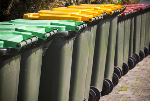 A row of large, dark green wheelie bins with brightly colored lids arranged along a pavement in an outdoor setting. The bins are positioned side by side, with the lids in yellow, green, and red, indicating different waste or rubbish categories. The lids are closed and appear to be made of durable plastic, with the black wheels visible at the base of each bin. The environment suggests a typical residential or communal rubbish storage area, possibly in or near Virginia Water, with a background of trees and natural greenery. The overall scene is well-lit, indicating daylight, and the ground surface is paved with concrete slabs, some of which are slightly mossy or weathered, reflecting the routine collection and disposal of waste managed by local rubbish removal services such as House Clearance Virginia Water.