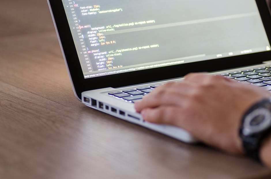 Close-up view of a person's right hand using a laptop with a silver metallic finish and a backlit keyboard, resting on a wooden surface. The laptop screen displays lines of code with syntax highlighti