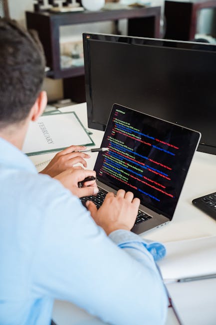 A person with short dark hair, dressed in a light blue shirt, sitting at a white desk while working on a silver laptop displaying lines of multicolored computer code. Next to the laptop, there is a bl